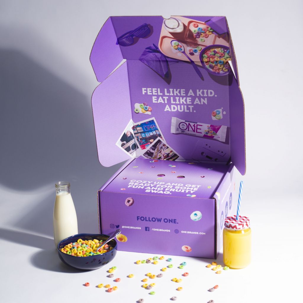 purple branded "One" box with brightly colored circled cereal on it and milk and a bowl of cereal next to it